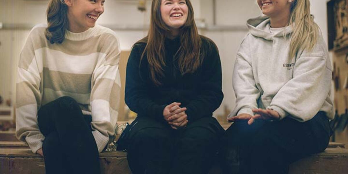 Group of three females sitting on workbench and smiling.