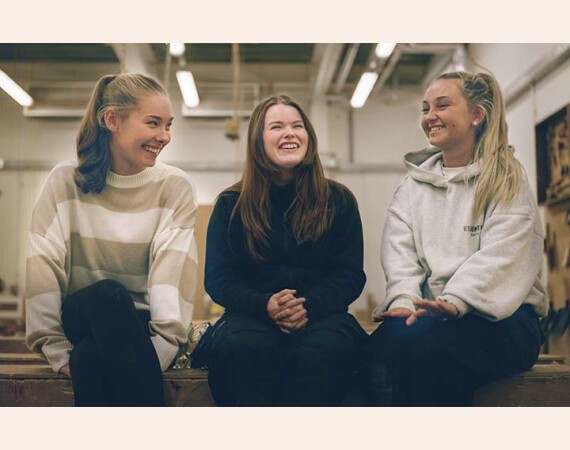 Group of three females sitting on workbench and smiling.