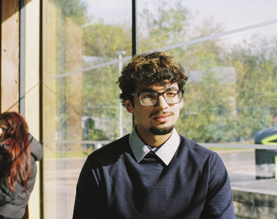 Male student with glasses sitting by window with female in background.