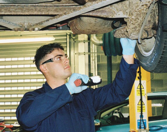 Automotive student looking at the underside of a car which is up on ramps. 