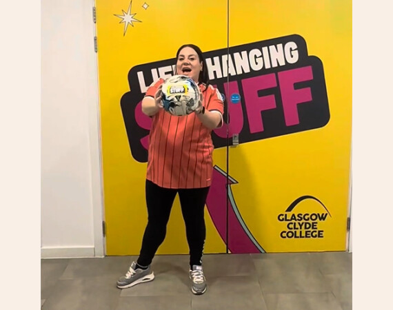 Image shows Anna Devitt holding a football in front of Life Changing Stuff sign at Glasgow Clyde College